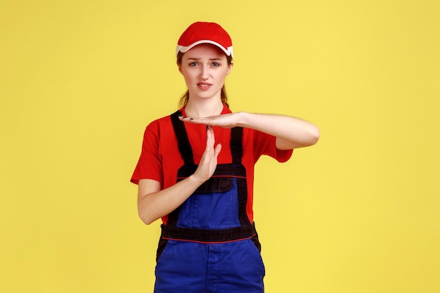 Retrato de mulher trabalhadora séria em pé olhando para a câmera mostrando gesto de tempo limite precisa de mais tempo vestindo macacão e boné vermelho Estúdio interno isolado em fundo amarelo