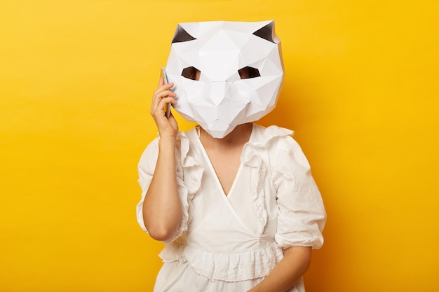 Retrato de mulher usando vestido e máscara de gato de papel posando isolado sobre fundo amarelo conversando no celular usando o celular