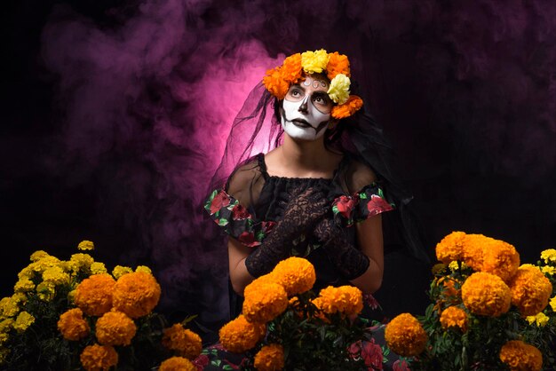 Foto retrato de mulher vestida de catrina rodeada de cempasuchil sobre fundo preto com fumaça