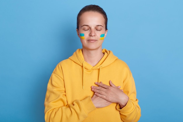 Retrato de mulher vestindo capuz com bandeira azul e amarela nas bochechas mantém as mãos no coração em pé com os olhos fechados pensando na guerra da Ucrânia contra a Rússia posando isolado sobre fundo azul