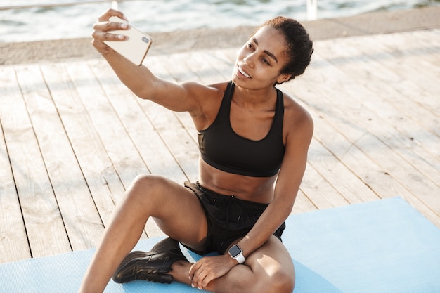 Retrato de mulher vestindo roupas esportivas tirando uma selfie no smartphone enquanto está sentada no tapete de finess à beira-mar pela manhã