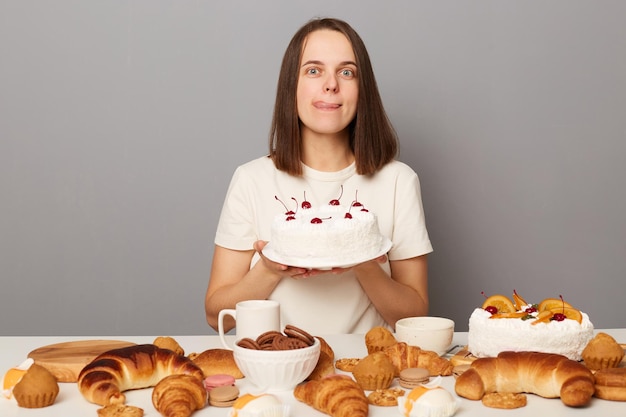 Retrato de mulher viciada em açúcar com fome com cabelo castanho vestindo camiseta branca sentada à mesa isolada sobre fundo cinza segurando o bolo mostrando a língua para fora