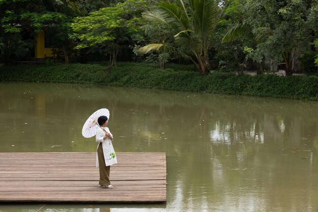 Retrato de mulher vietnamita em vestido ao dai segurando um guarda-chuva de papel perto do lago