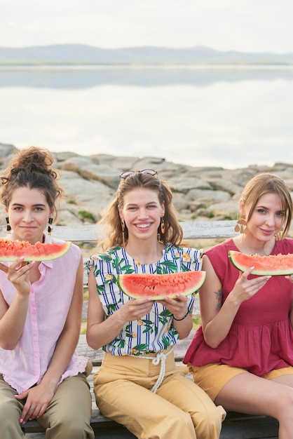 Retrato de mulheres jovens e bonitas segurando fatias de melancia e sorrindo alegremente para a câmera enquanto está sentado no fundo do lindo lago