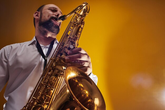 Retrato de músico profissional saxofonista homem de camisa branca toca música jazz no saxofone, fundo amarelo em um estúdio fotográfico, vista inferior