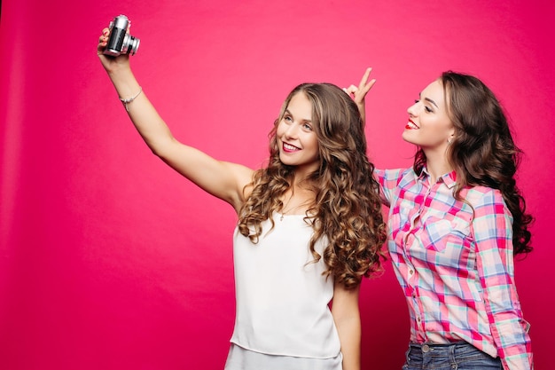 Retrato de namoradas atraentes felizes com longos penteados ondulados tirando auto-retrato via câmera de filme antigo contra fundo vermelho Morena de camisa xadrez fazendo orelhas de coelho para sua amiga
