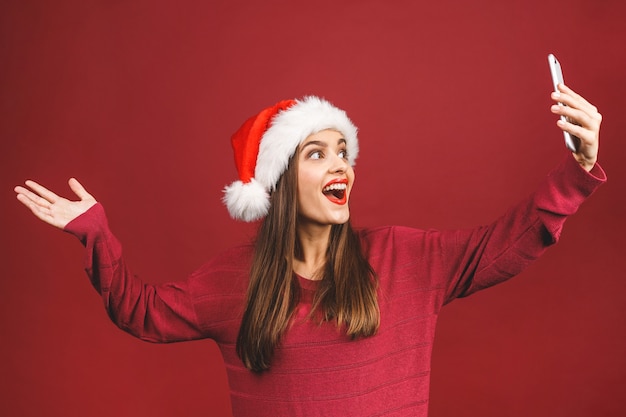 Retrato de Natal da linda menina com chapéu de Papai Noel, sorrindo e fazendo selfie no celular