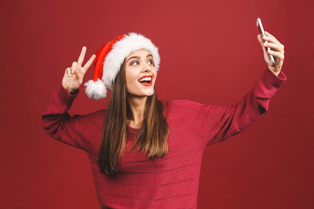 Retrato de Natal de uma linda garota com chapéu de Papai Noel, sorrindo e fazendo selfie no celular