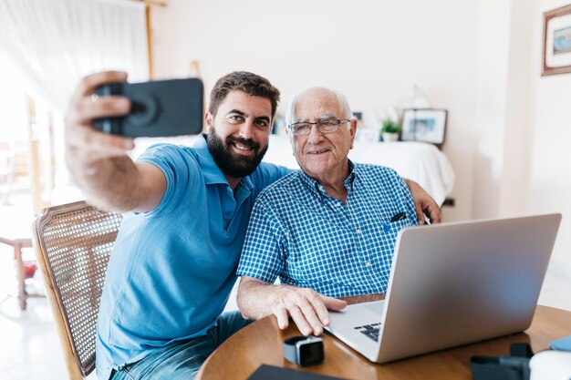 Retrato de neto adulto e seu avô tirando selfie com smartphone em casa