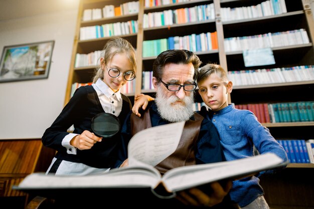 retrato de netos adolescentes atraentes e experiente avô barbudo sênior que passar algum tempo juntos na biblioteca na leitura de livro interessante. Estante no fundo