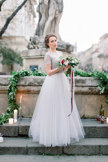 Retrato de noiva linda no vestido de casamento incrível segurando buquê macio em pé ao ar livre nas escadas. Antigo centro da cidade, Lviv, Ucrânia