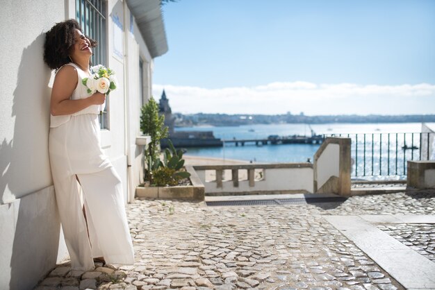 Retrato de noiva rindo com buquê. Vista lateral de uma mulher encaracolada em um vestido branco em pé na parede, sorrindo