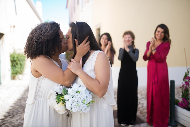 Retrato de noivas se beijando fofos. Duas jovens se abraçando, se beijando ternamente. Convidadas rindo e aplaudindo ao fundo