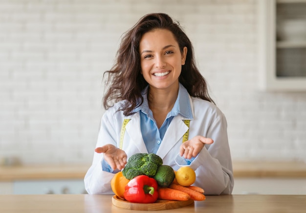 Retrato de nutricionista alegre com jaleco apontando para frutas e legumes frescos na mesa sorrindo