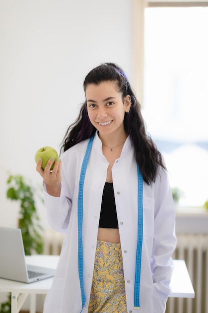 Retrato de nutricionista feminina sorridente de jaleco branco com maçã fresca na mão, ficando no escritório