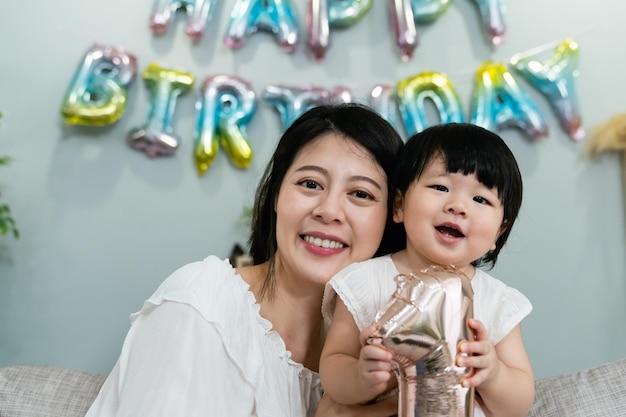 retrato de ombro de uma doce mãe asiática e um bebê de um ano posando juntos e sorrindo para a câmera em um interior de casa festivo com decoração de aniversário