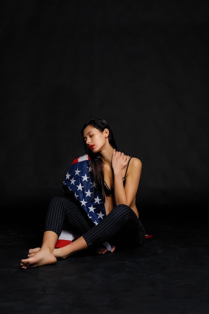 Retrato de orgulhosa atleta envolto na bandeira americana contra fundo preto. Mulher jovem e musculosa, olhando com confiança para a câmera.