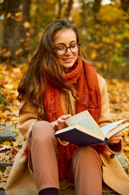 Retrato de outono aproximado de uma jovem inteligente de óculos e roupas quentes lendo livro ao ar livre