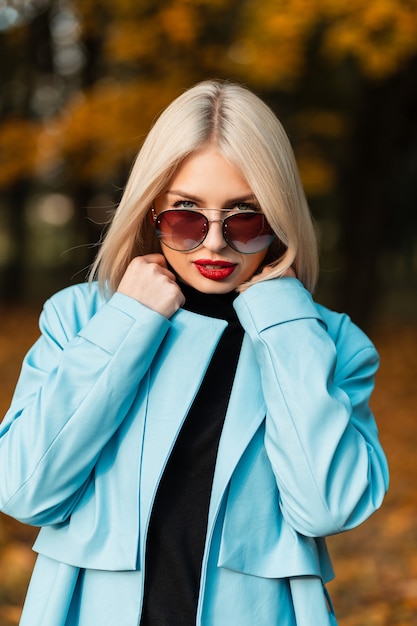 Retrato de outono de uma linda mulher loira com lábios vermelhos em roupas da moda com óculos de sol no fundo de um parque de outono com folhagem de cor brilhante