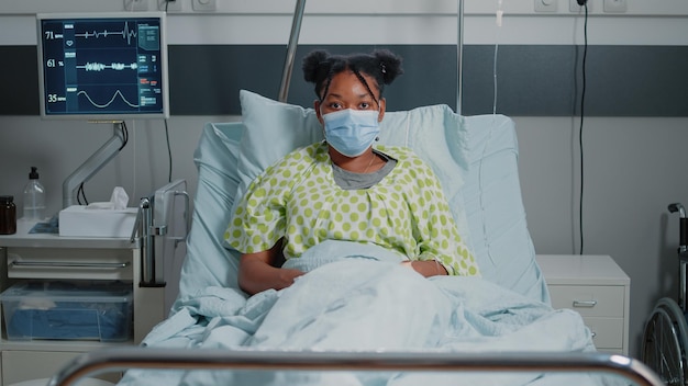 Retrato de paciente afro-americano usando máscara facial e esperando para receber assistência médica e tratamento contra doenças. mulher doente sentada na enfermaria do hospital durante a pandemia.