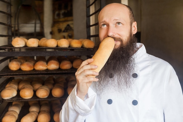 Retrato de padeiro adulto jovem alegre com barba longa em uniforme branco permanente em sua fabricação, cheirando com prazer pão fresco e desfrutando. olhando para cima, interior, conceito de profissão