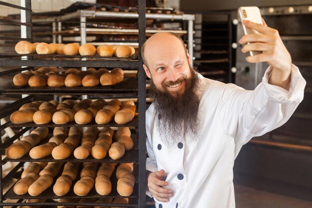 Retrato de padeiro de blogueiro adulto jovem alegre com barba longa em uniforme branco em pé na fábrica e fazendo selfie nas prateleiras com fundo de pão fresco, piscando, interior, conceito de profissão