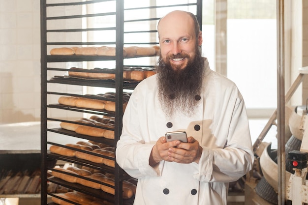 Retrato de padeiro de empresário adulto jovem satisfeito com barba longa em uniforme branco em pé na padaria e ter pedido on-line por telefone, usando o telefone com sorriso interno, olhando para a câmera