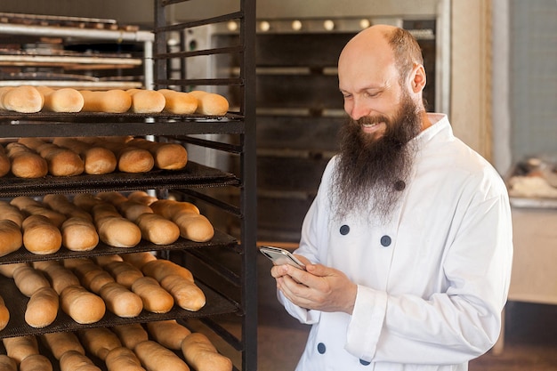 Retrato de padeiro de empresário adulto jovem satisfeito com barba longa em uniforme branco em pé na padaria e ter pedido on-line por telefone usando telefone com sorriso de dentes Conceito de profissão interna