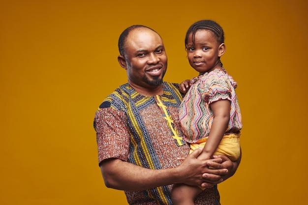 Retrato de pai africano segurando sua filhinha e sorrindo para a câmera em pé contra ba amarelo