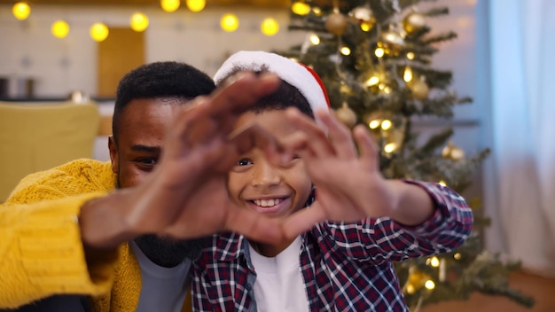 Retrato de pai afro-americano e filho gesticulando coração com as mãos olhando para a câmera perto da árvore de natal