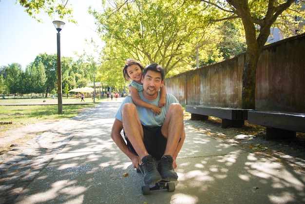 Retrato de pai asiático e filho se divertindo com skate. Homem sorridente sentado no skate e filha em pé atrás dele no beco, ambos olhando para a câmera. Descanso ativo e conceito de infância feliz