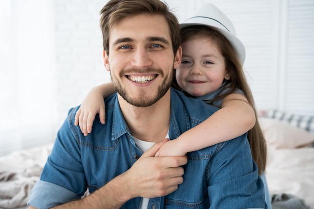 Foto retrato de pai bonito e sua linda filha abraçando olhando para a câmera e sorrindo enquanto está sentado no sofá em casa