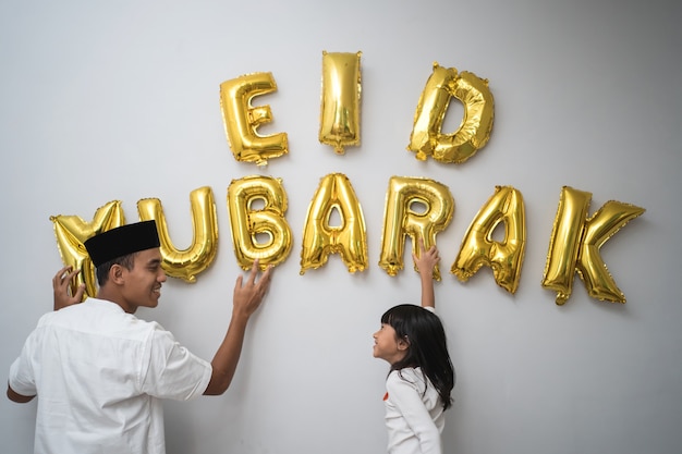 Foto retrato de pai e filha muçulmanos decorando uma carta de eid mubarak feita de uma decoração de balão contra a parede em casa