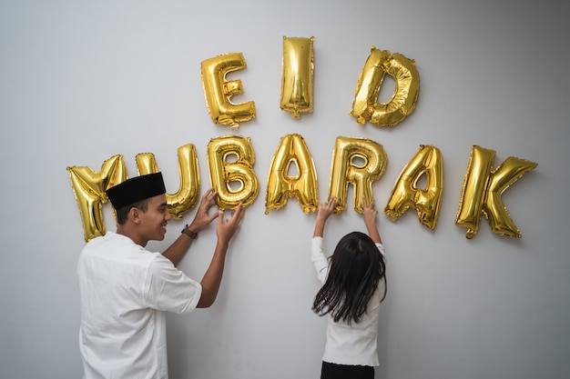 Retrato de pai e filha muçulmanos decorando uma carta de eid mubarak feita de uma decoração de balão contra a parede em casa