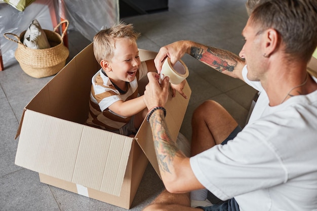 Retrato de pai feliz brincando com menino bonito em caixa de papelão enquanto faz as malas para a família mudar de casa