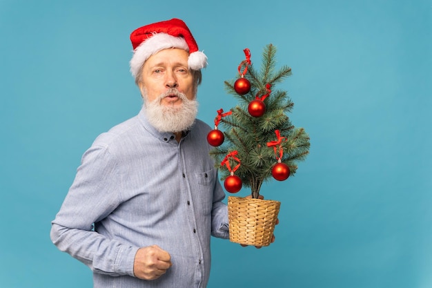 Retrato de Papai Noel surpreso segura árvore de natal em fundo azul com emoções de espaço de cópia e conceito de férias de inverno