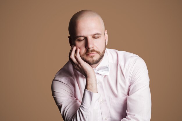 Retrato de pensar bonito homem careca barbudo na camisa rosa claro e laço branco, sentado na cadeira, tocando seu rosto com a mão e os olhos fechados. tiro do estúdio interno, isolado no fundo marrom.