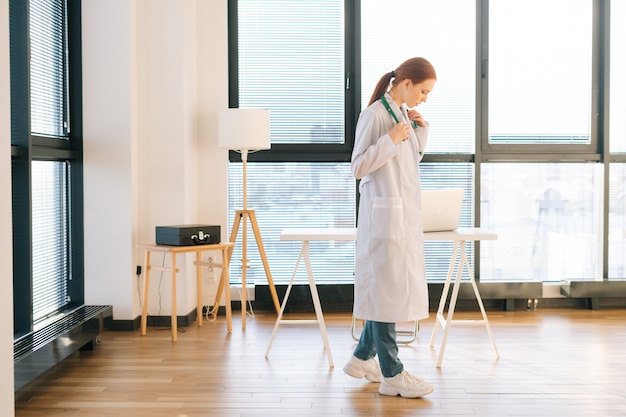 Retrato de pensar médica em pé de jaleco branco no fundo da janela em um dia ensolarado no escritório da clínica médica leve.