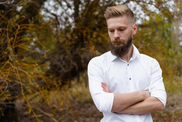 Retrato de pensativo barbudo jovem atraente em camisa branca, olhando para um parque de outono em outubro.