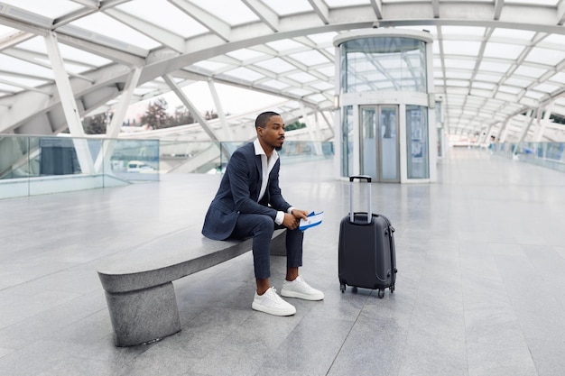 Retrato de pensativo jovem empresário negro sentado no banco no aeroporto