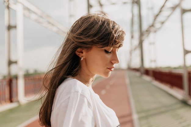 Retrato de perfil de uma linda mulher com sorriso bonito e olhos fechados está descansando do lado de fora da cidade Senhora morena feliz com roupa elegante está indo na ponte à luz do sol