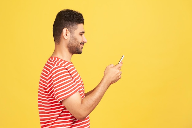 Retrato de perfil homem barbudo feliz em mensagens de texto de t-shirt listrada vermelha no smartphone, tendo conversa online em redes sociais. Tiro de estúdio interior isolado em fundo amarelo