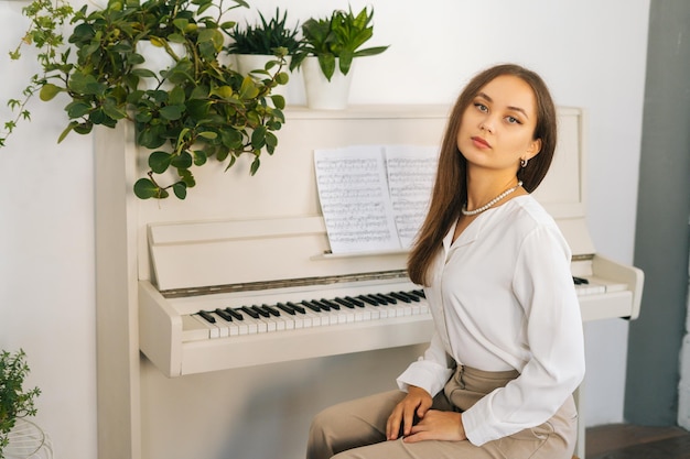 Retrato de pianista jovem confiante olhando para a câmera sentada no piano branco clássico em sala de aula