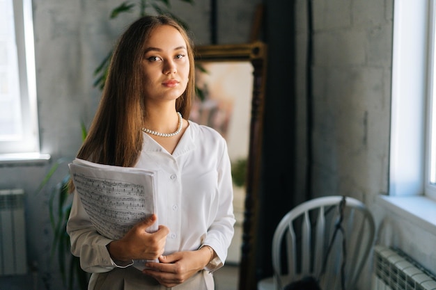 Retrato de pianista muito jovem segurando nas mãos partitura com notas musicais na sala de aula escura, olhando para a câmera