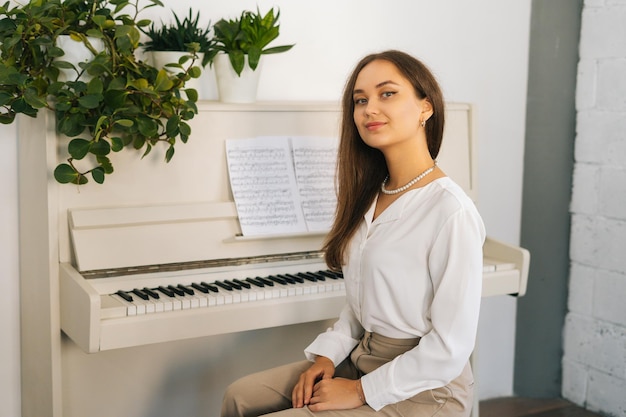 Retrato de pianista sorridente jovem olhando para a câmera sentada no piano branco clássico em sala de aula