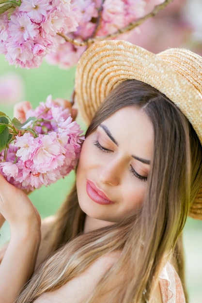 Foto retrato de primavera de uma jovem linda em flores cor de rosa jovem linda modelo com cabelo comprido em um chapéu de vime e um vestido leve perto das flores de cerejeira