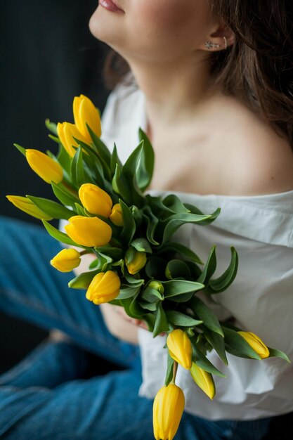 Retrato de primavera de uma jovem mulher bonita com olhos azuis e cabelos cacheados com buquê de tulipas amarelas em fundo preto Modelo feliz em jeans e camisa branca dia das mães