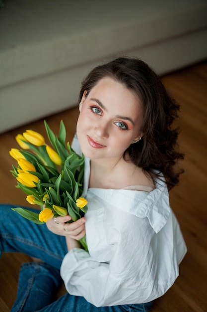 Retrato de primavera de uma jovem mulher bonita com olhos azuis e cabelos cacheados com buquê de tulipas amarelas no interior de casa modelo feliz em jeans e camisa branca dia das mães