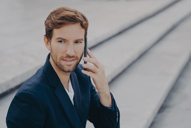 Retrato de profissional de negócios atraente confiante fazendo ligação, falando no smartphone, conversando de negócios com o parceiro, desviando o olhar enquanto está sentado do lado de fora nas escadas de pedra no centro da cidade