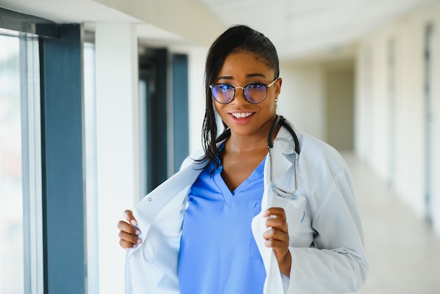 Retrato de profissional de saúde feminino confiante, sorridente e amigável com jaleco, braços cruzados segurando os óculos. Fundo de clínica hospitalar isolado.